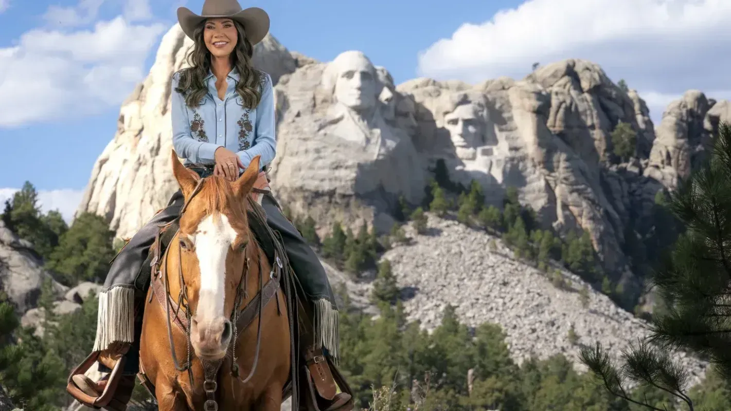 A woman wearing a cowboy hat and a blue shirt is riding a brown horse in front of Mount Rushmore, with the faces of four U.S. presidents carved into the rocky landscape.